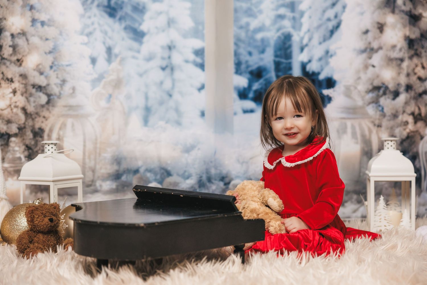 A child in a red holiday dress sits on a white winter wonderland set with lanterns and snowy decorations.