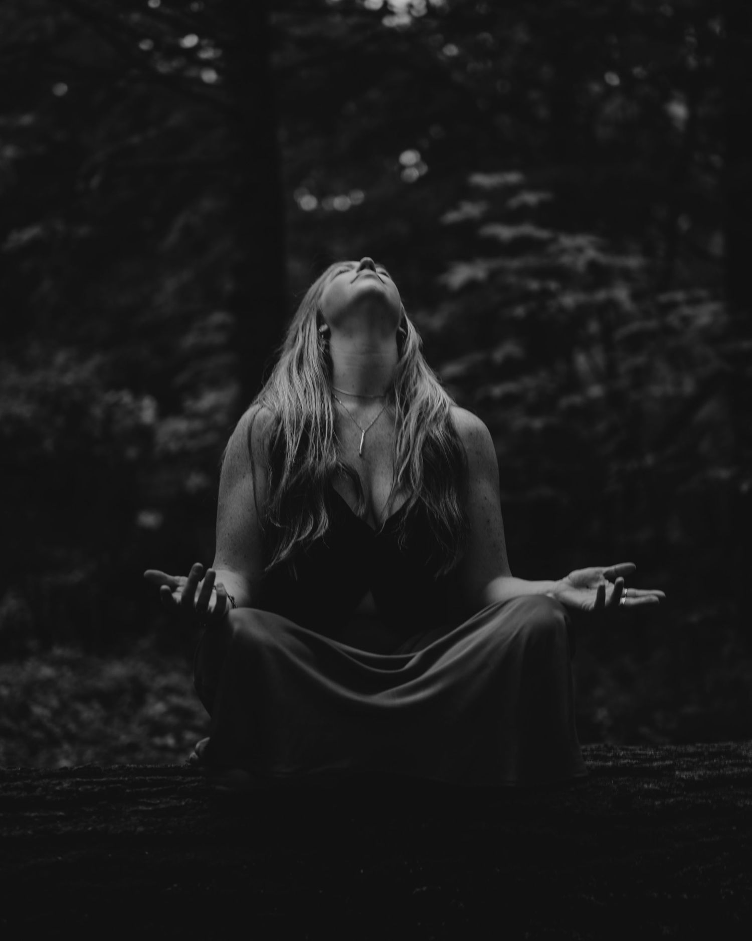 A figure in a dark dress sitting in meditation pose against a moody forest background in black and white.