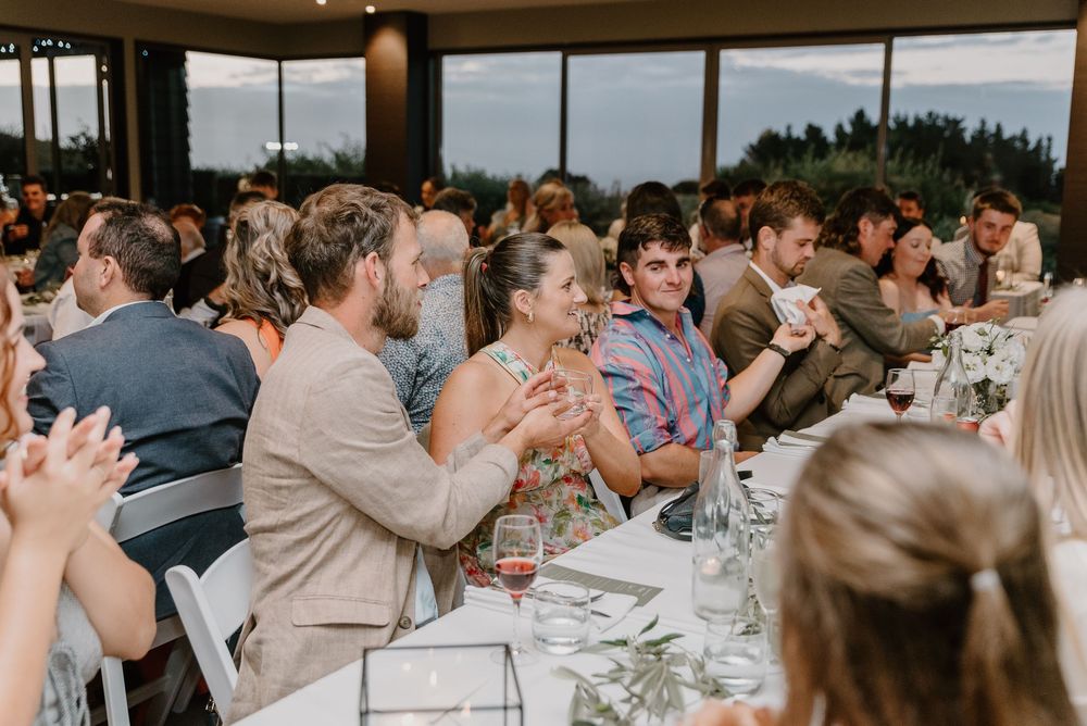 Wedding guests clap and celebrate during a reception dinner at a venue with panoramic windows.