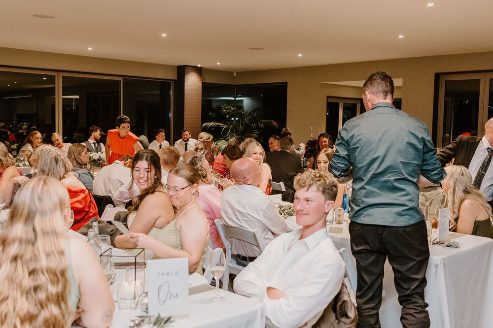 Reception guests seated at white-clothed tables listen during an indoor evening event at an elegant venue.