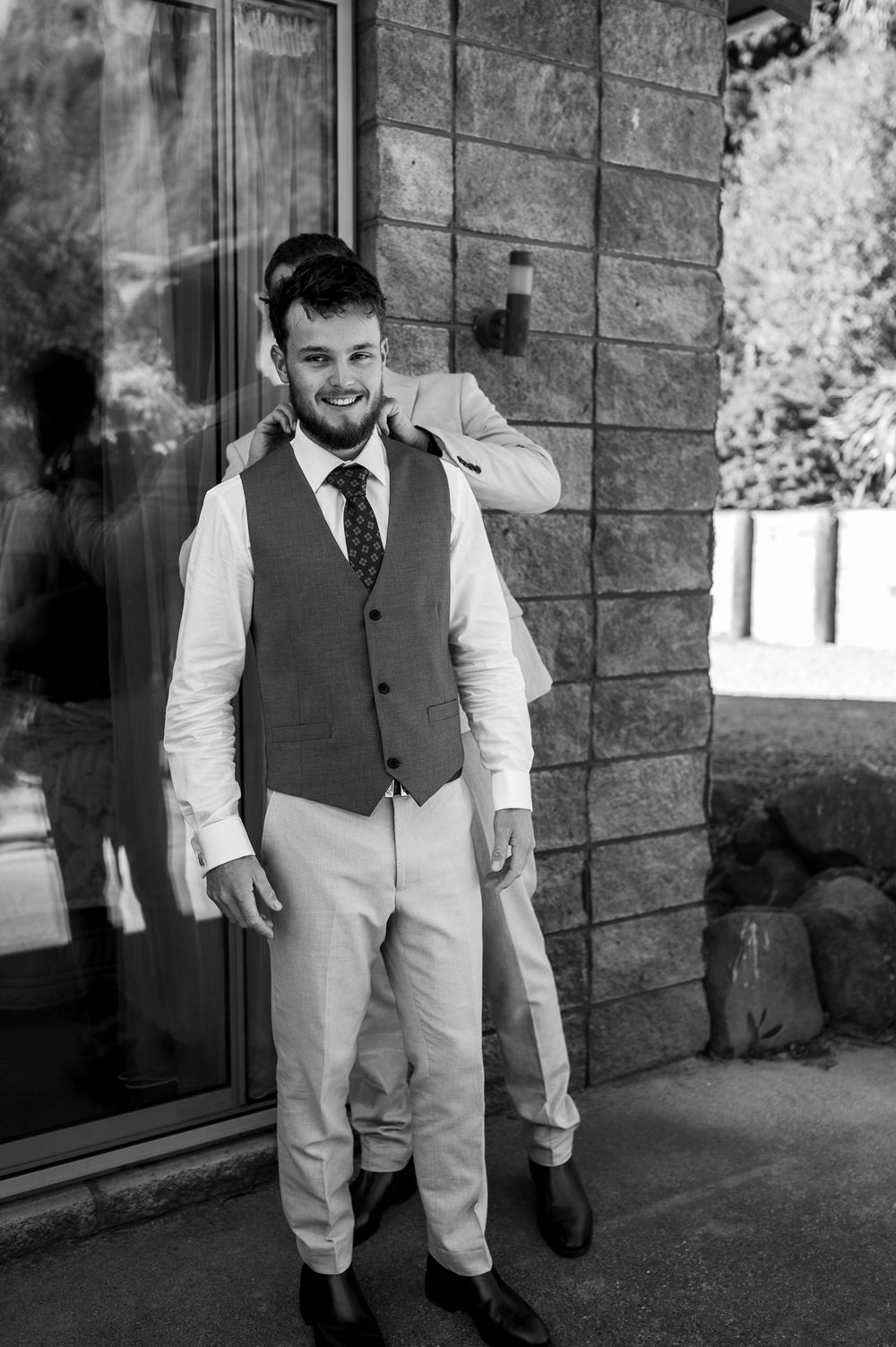 A groom in a formal vest and tie stands by a stone wall in a black and white photograph.