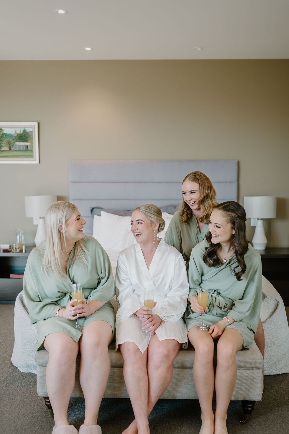 Four friends in matching sage green robes share joyful moments while sitting together on a bed in a hotel room.
