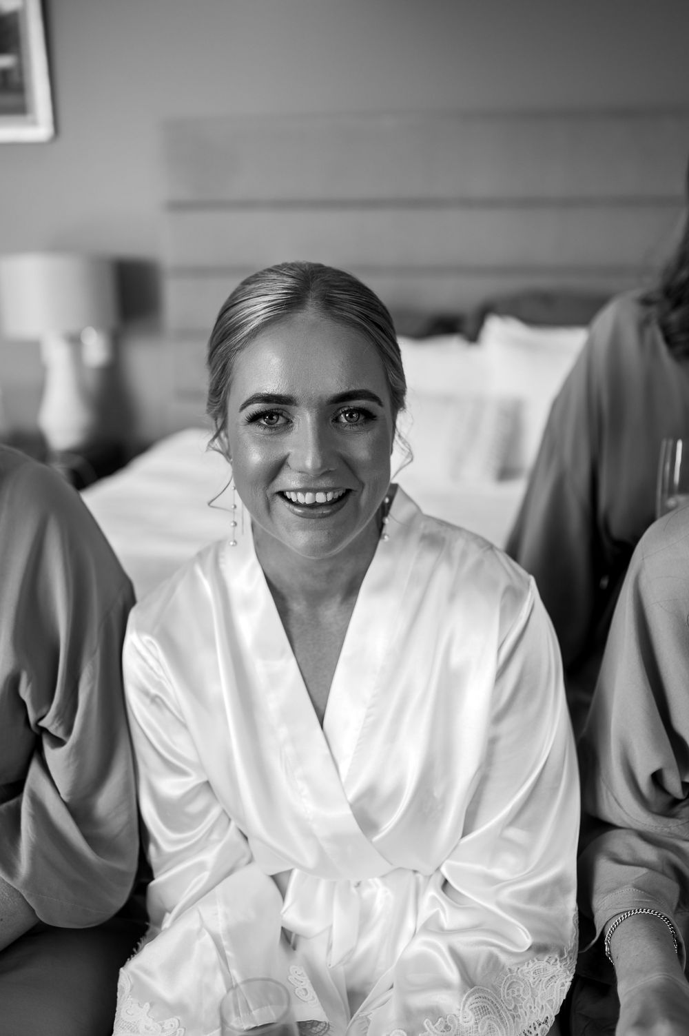 A woman in a white silk robe smiles brightly while getting ready on a wedding day morning.