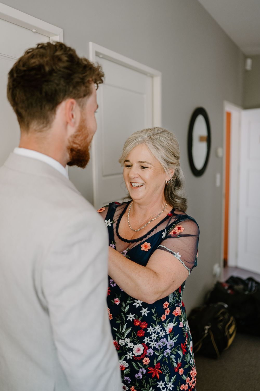 Two people share a joyful moment in a hallway, one wearing a white suit and another in a floral print mesh dress.