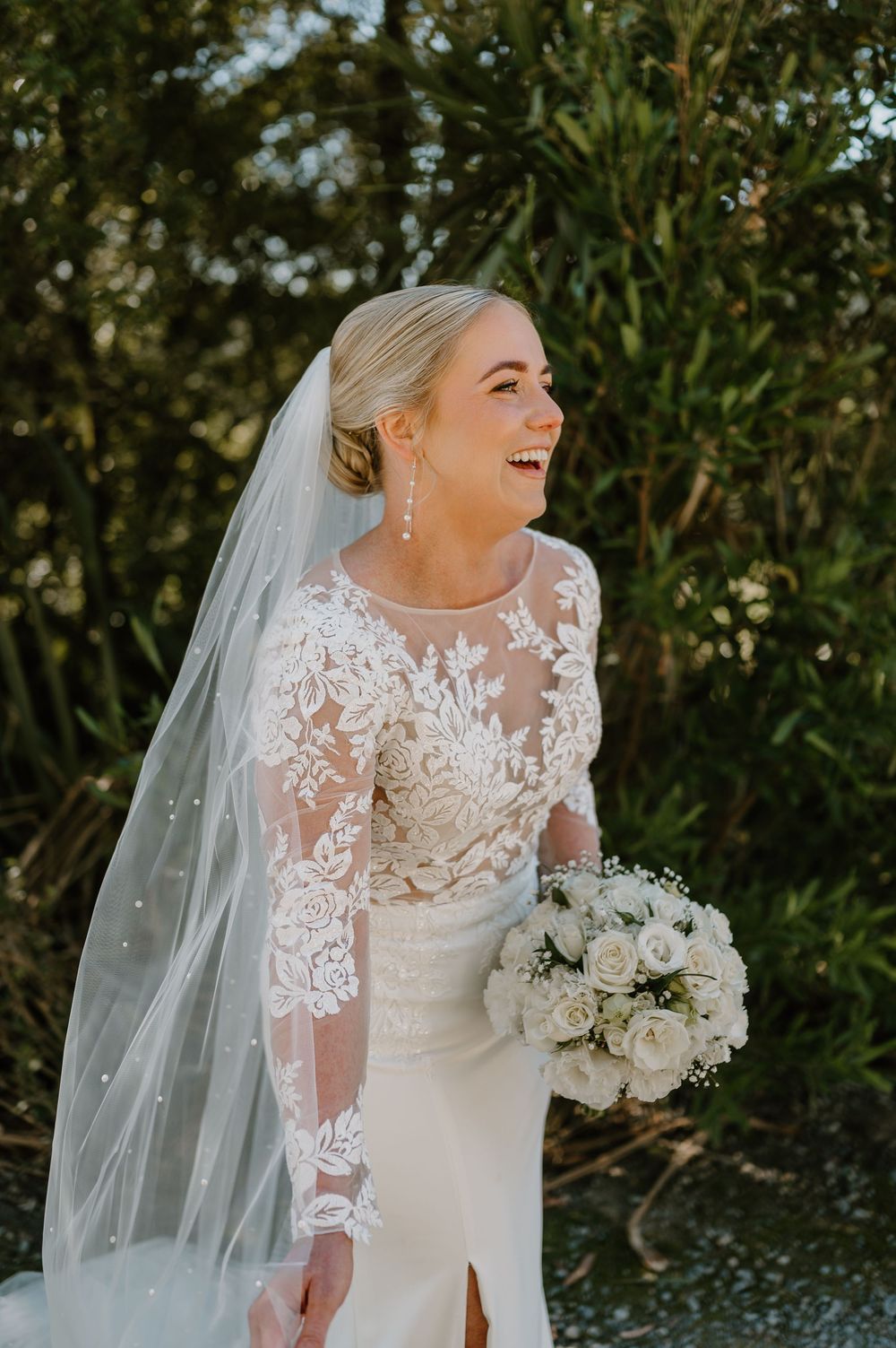 A bride in a lace sleeve wedding gown and veil holds a white bouquet while laughing outdoors in natural light.