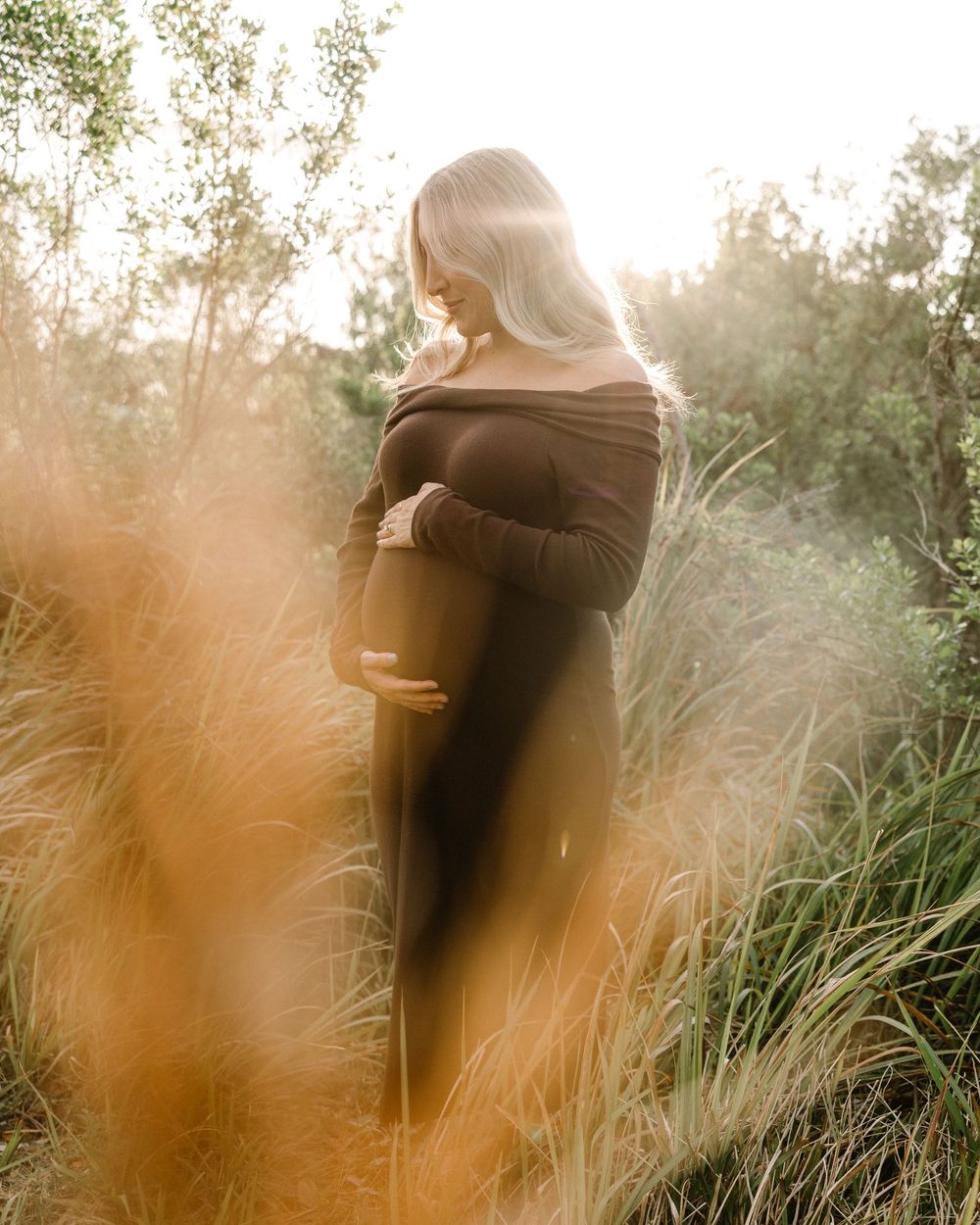 Artistic maternity photo through tall grass at golden hour.