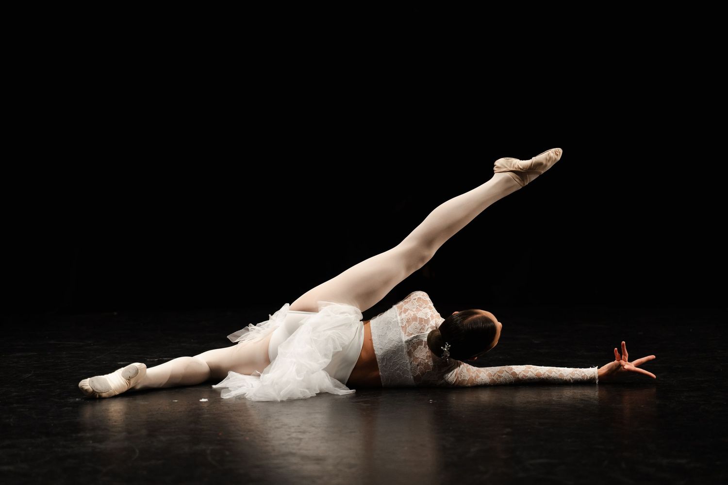 Ballet dancer in white costume performing graceful floor movements on dark stage under dramatic lighting.