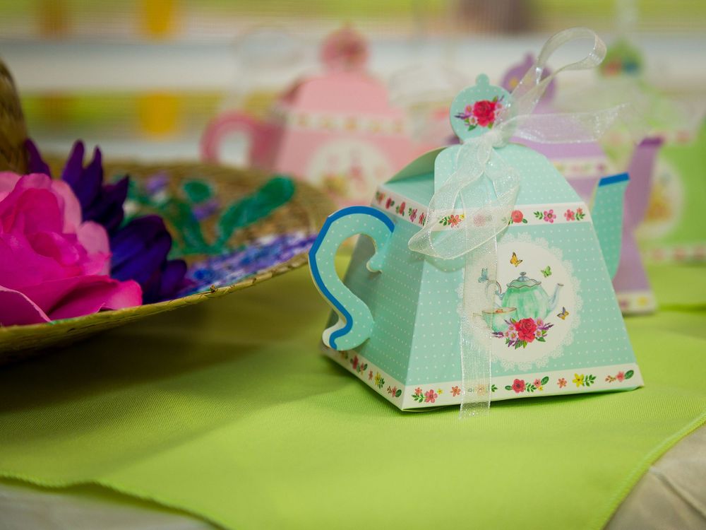 Decorative mint green paper teapot favor box with delicate floral designs and gold trim sits on lime green surface.