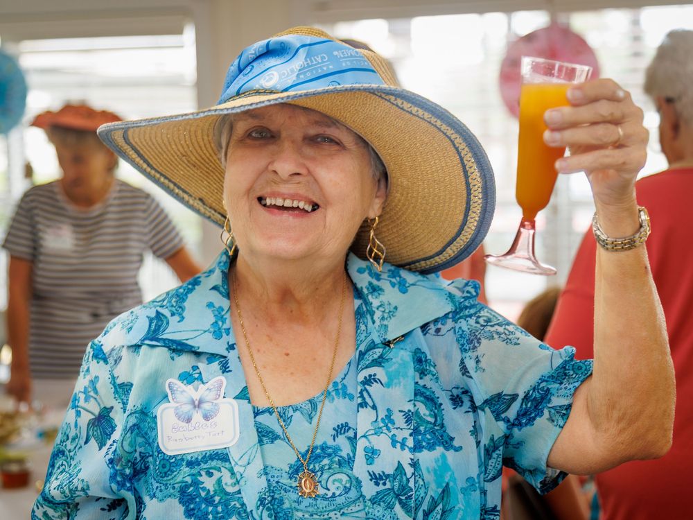 Senior wearing a blue floral top and wide-brimmed sun hat raises a drink while enjoying an outdoor gathering.