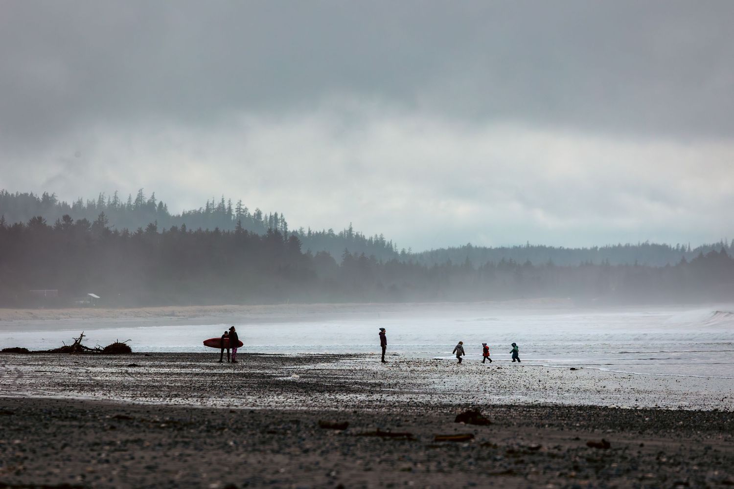 Silhouettes of people walking on a misty beach north of Masset on Haida Gwaii with forested hills in the background during an overcast day.