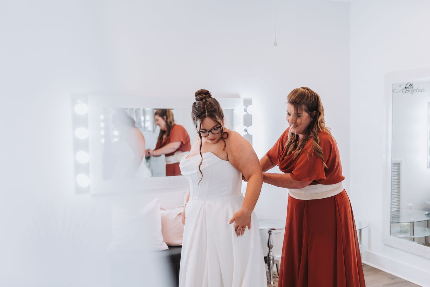 A group in a bright white dressing room helps someone put on a wedding dress during preparation.