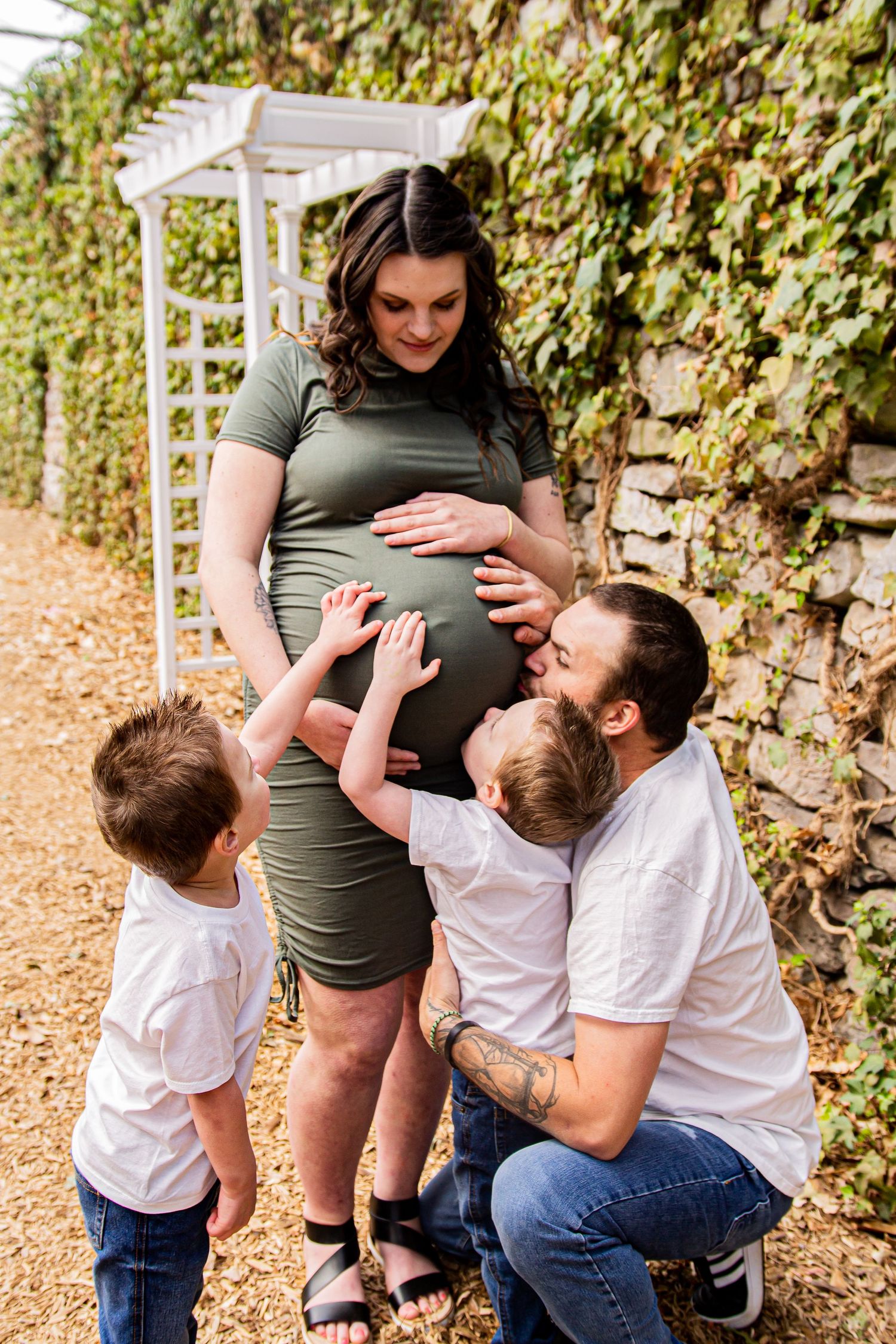 Expectant family sharing tender moment in garden setting with ivy-covered wall.