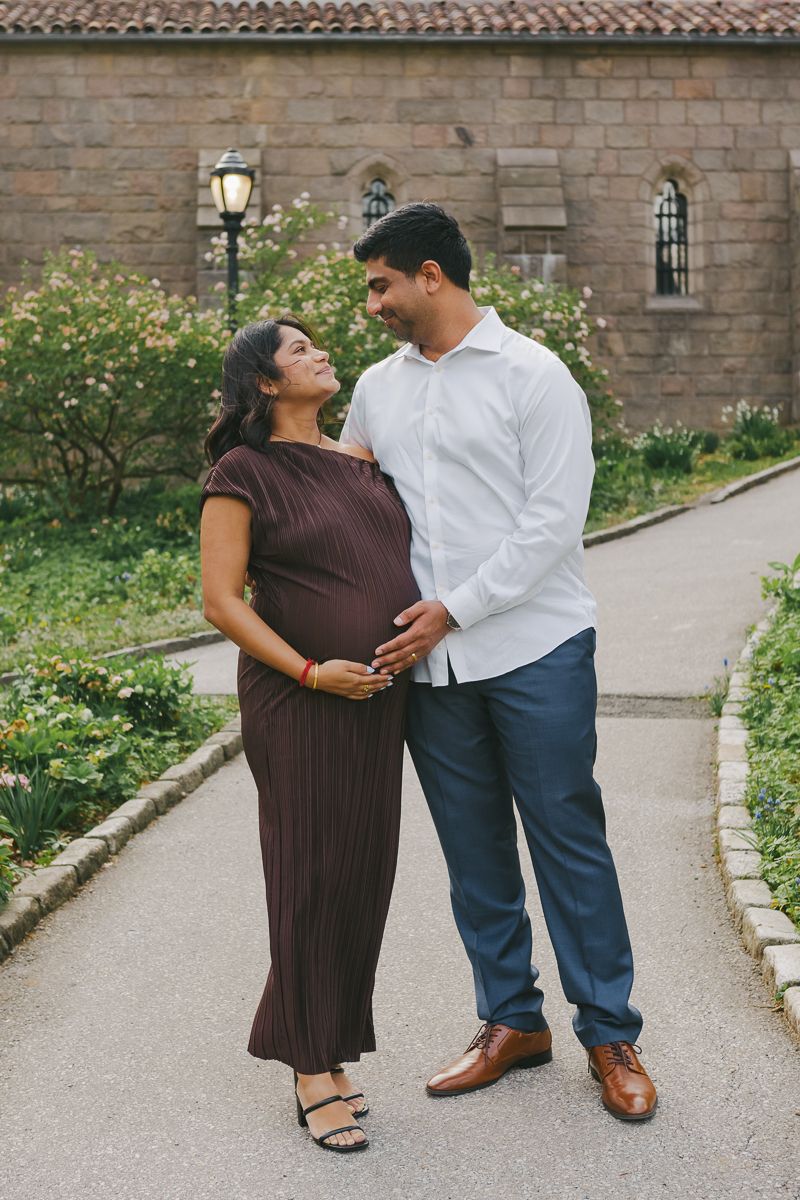 Expecting couple gazing at each other, surrounded by the golden glow of sunset at Fort Tryon Park, by NYC maternity photographer Paula B