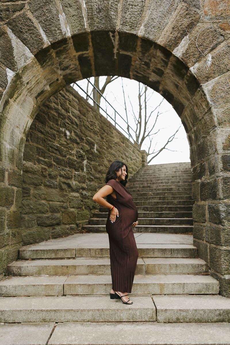 Mom-to-be standing gracefully under the stone arch, embraced by soft golden light, by NYC maternity photographer Paula Brearley.