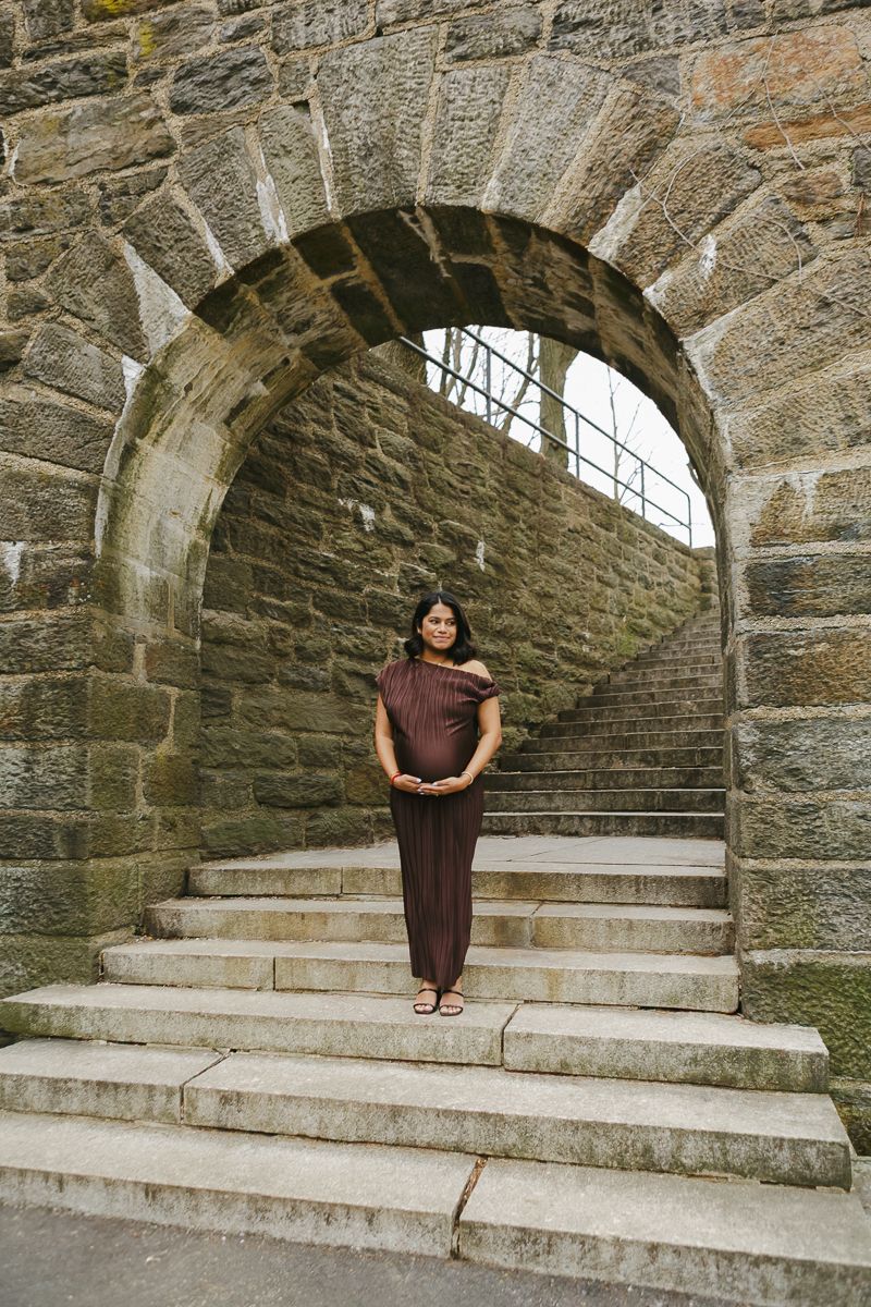 Expecting mother cradling her baby bump, framed under a dramatic stone archway bathed in golden sunlight by Paula Brearley NYC maternity