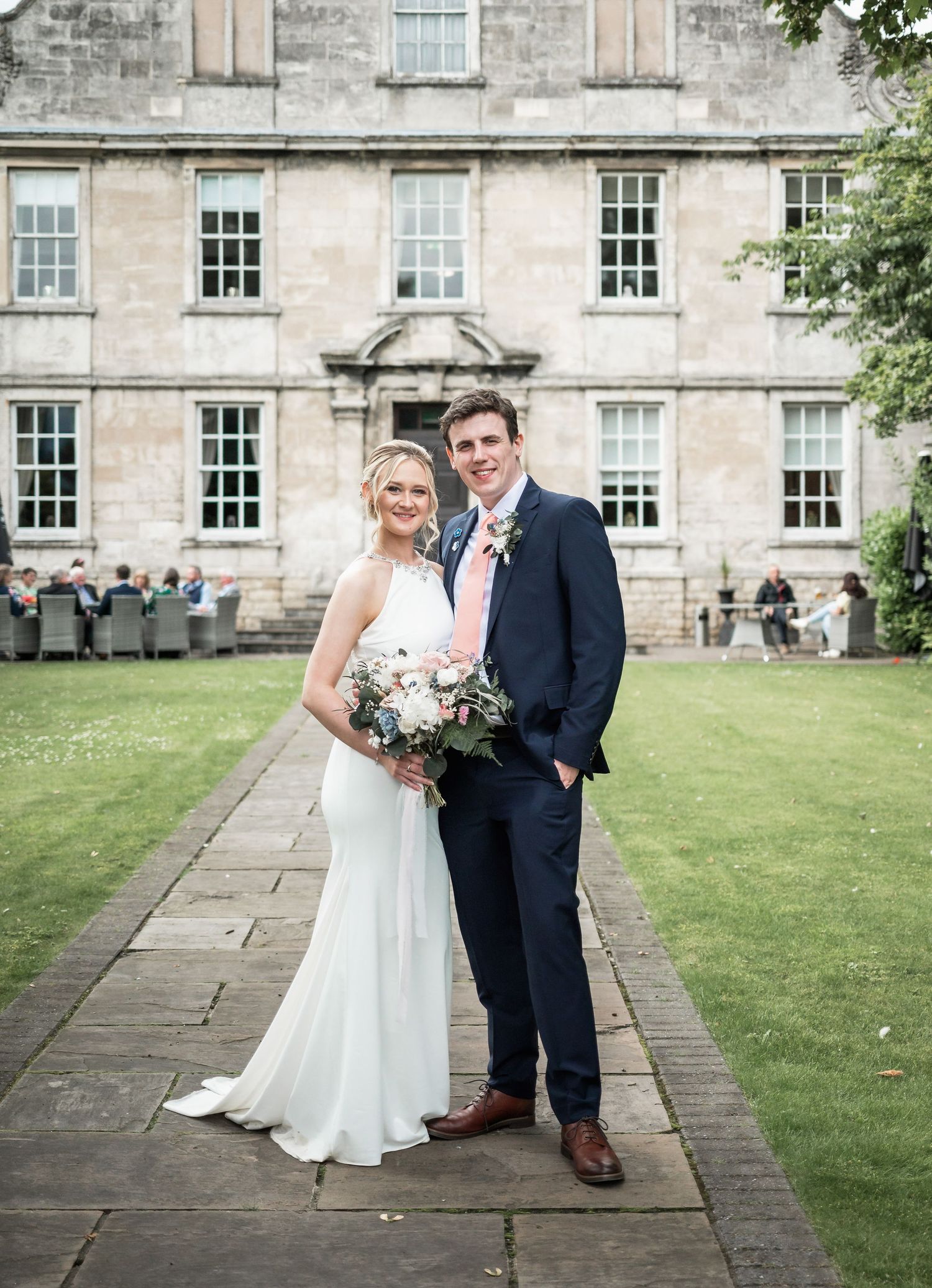 Happy newlyweds embrace in a garden setting with lush greenery and a grand stone building in the background.