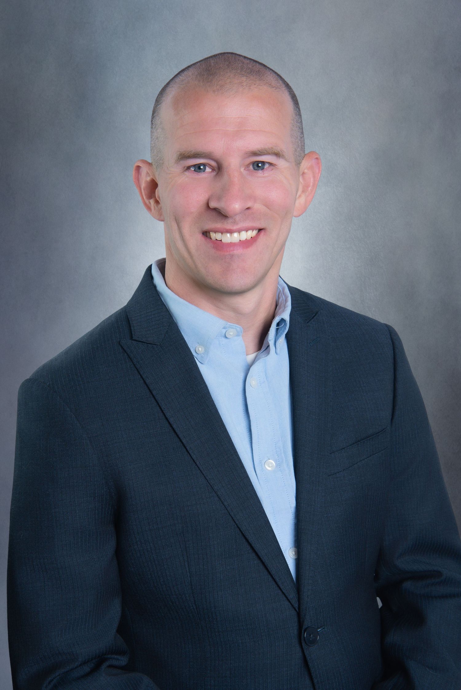 Professional headshot of someone in a dark blazer and light blue button-down shirt against a gray backdrop.