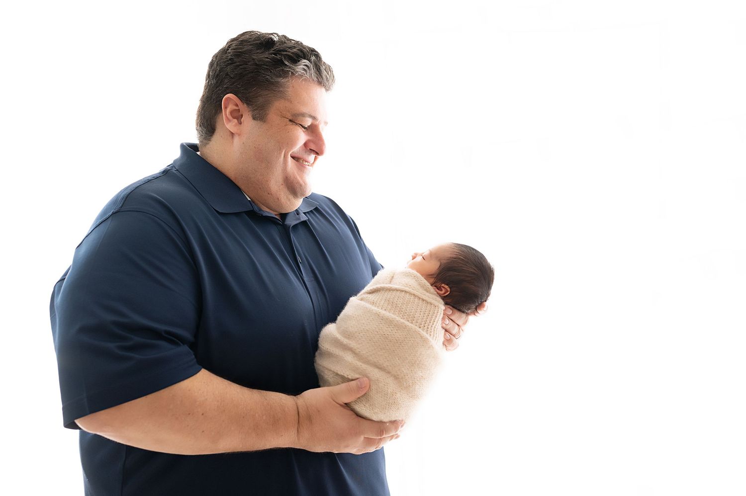 Proud parent in navy polo shirt tenderly holding swaddled newborn against white background.