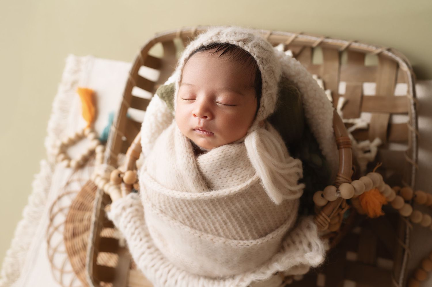 Newborn baby sleeping peacefully in beige knit wrap on woven basket prop.