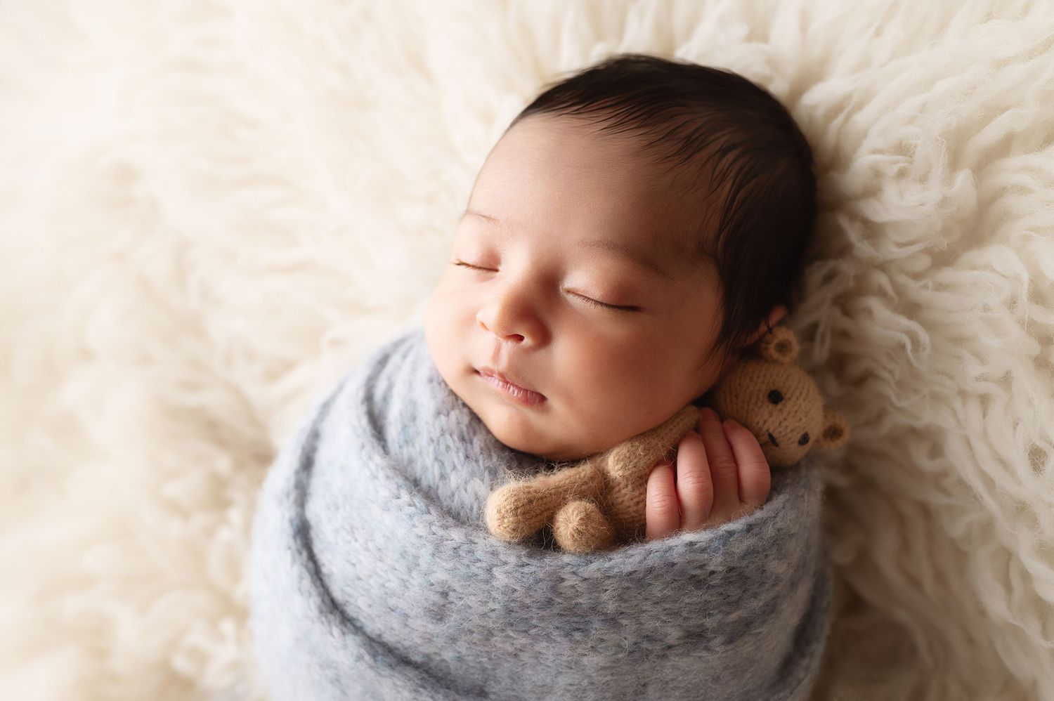 Peaceful newborn baby swaddled in soft grey blanket resting on cream-colored fur backdrop.
