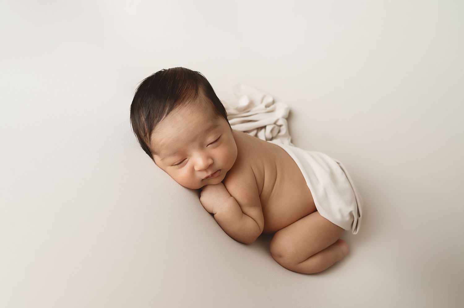 Sleeping newborn baby wrapped in white fabric lying on neutral background during professional photo session.