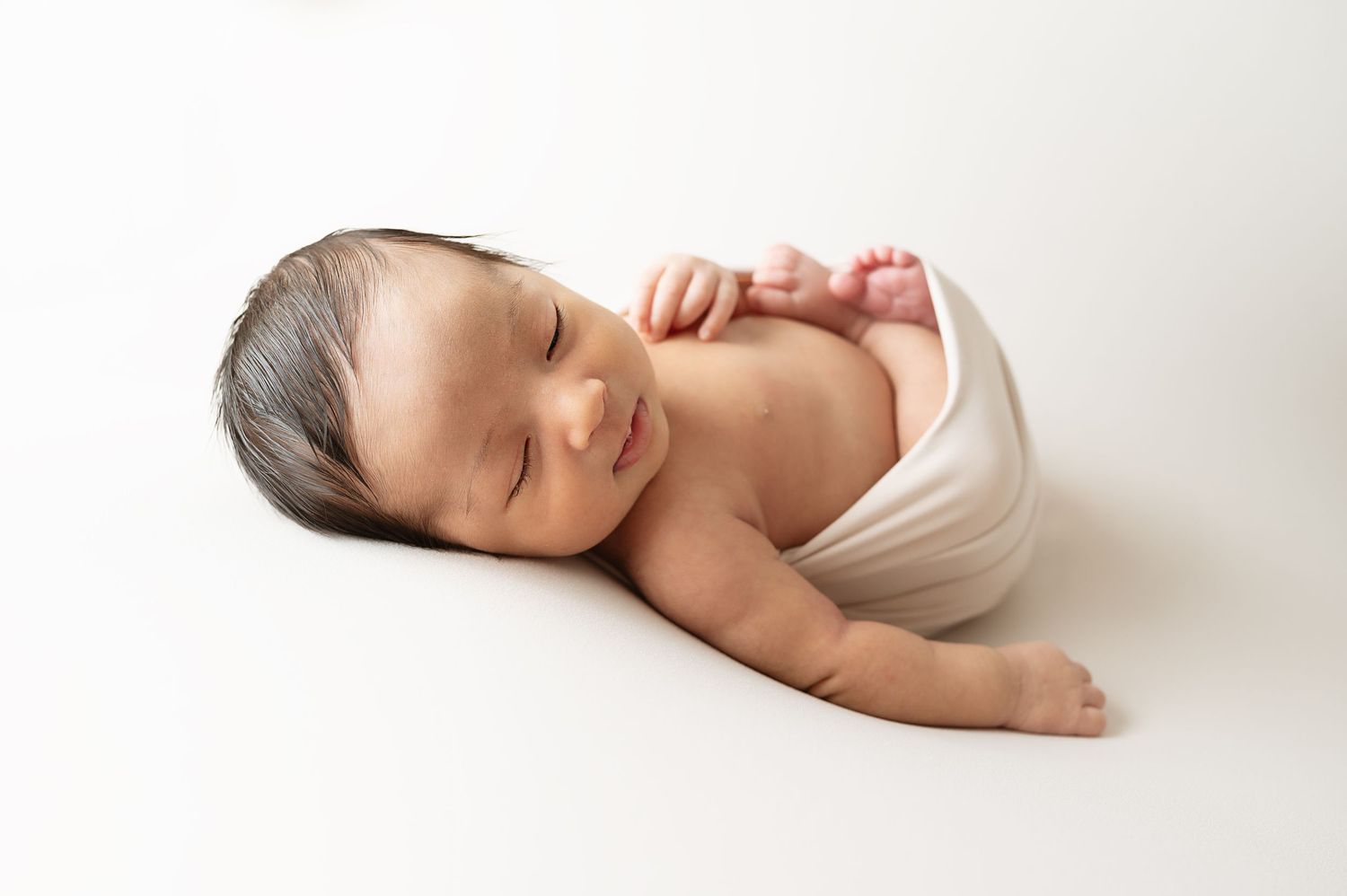 Sleeping newborn wrapped in white fabric lying on simple white background during portrait session.