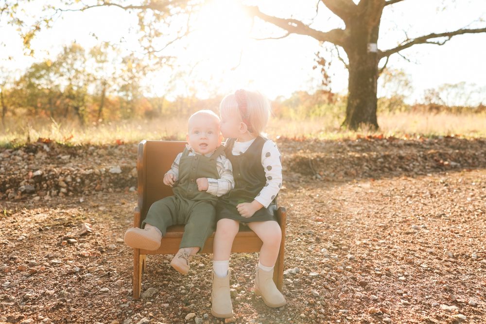 Two toddlers sitting together on wooden chair outdoors during sunset.