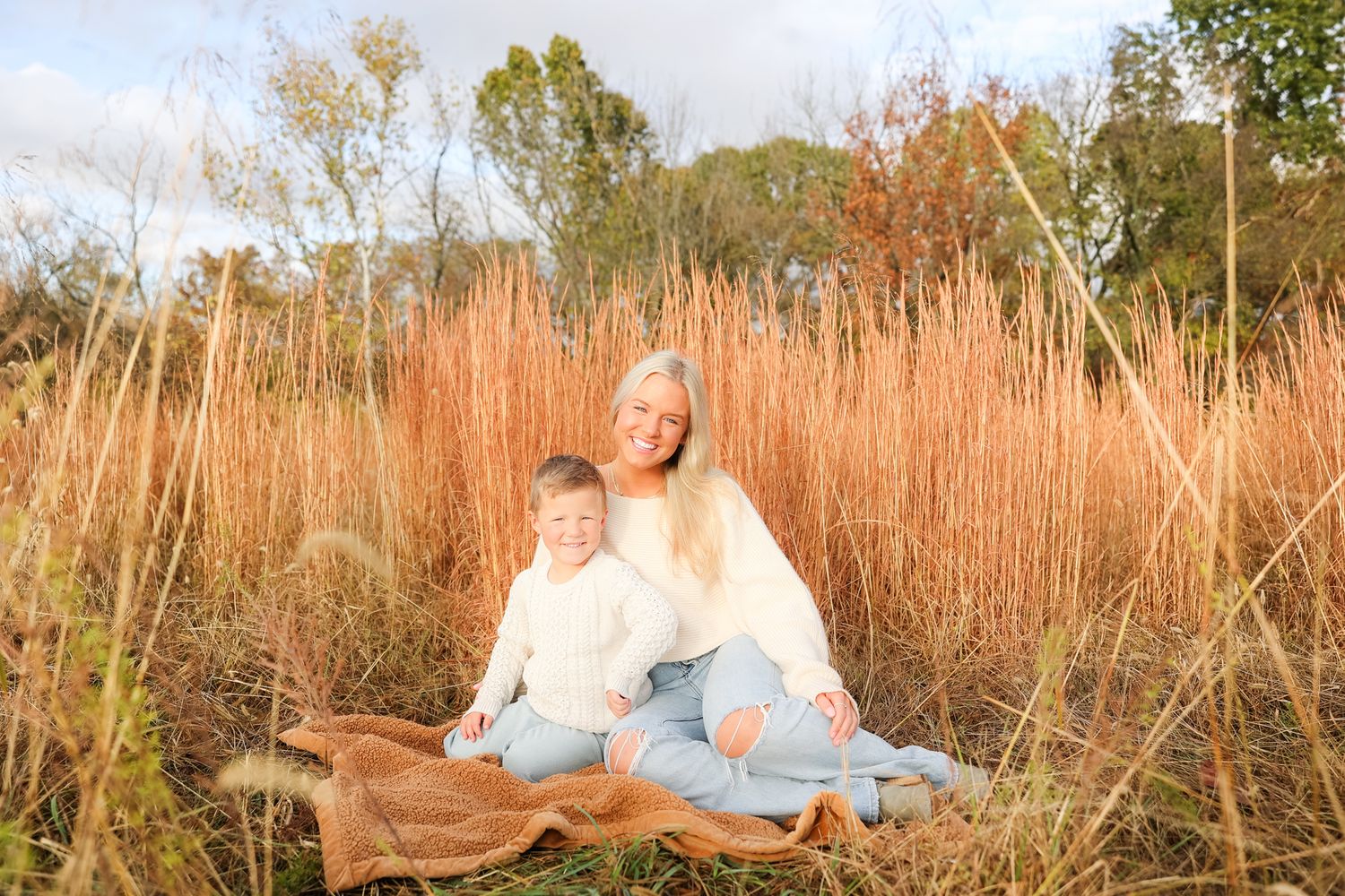Mother and son sitting on blanket in field of tall golden grasses during autumn.