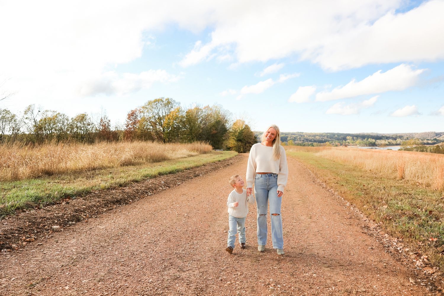 Mother and son walk hand-in-hand down a dirt road surrounded by tall grass.