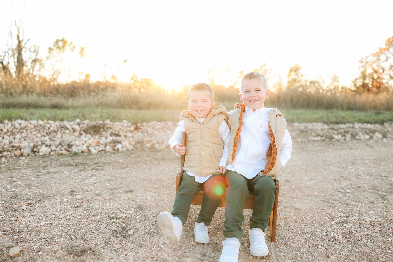 Two children sit together on a wooden bench in a field at sunset.