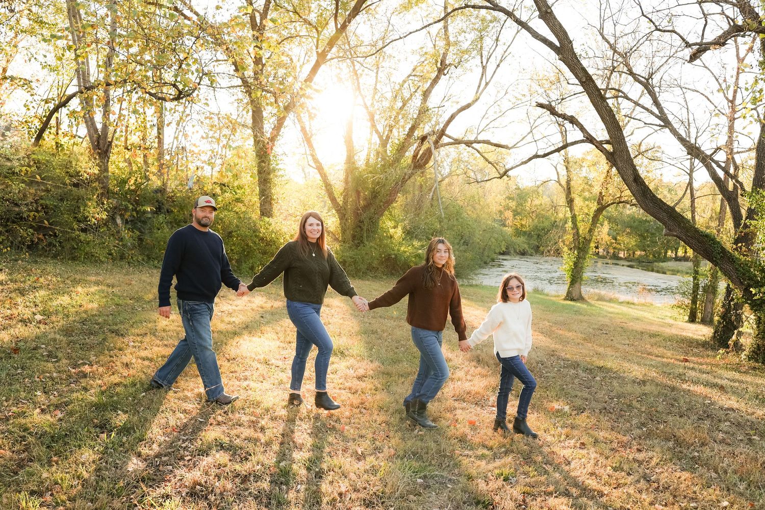 Family holds hands while walking through sun-dappled autumn park.