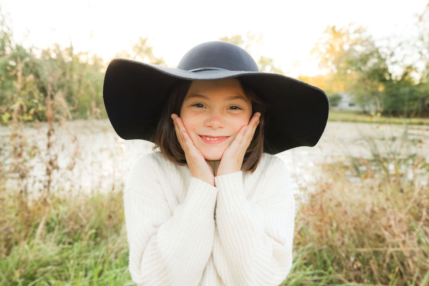 Child wearing a large black sun hat and white sweater smiling in a sunny outdoor field.