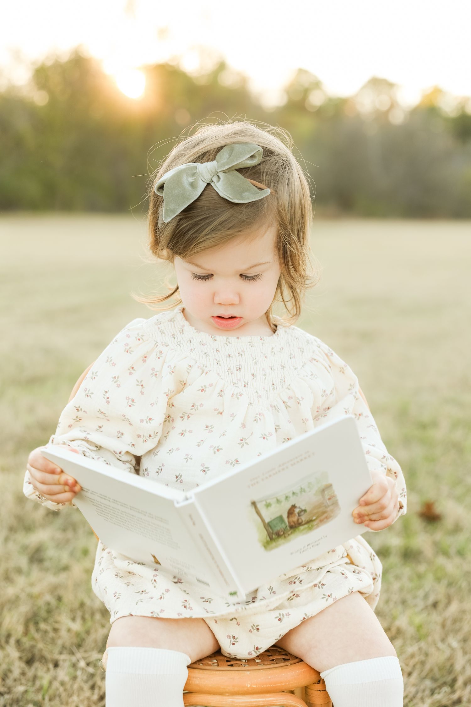 A small child in a white floral dress reads a book while sitting outdoors during golden hour sunset.