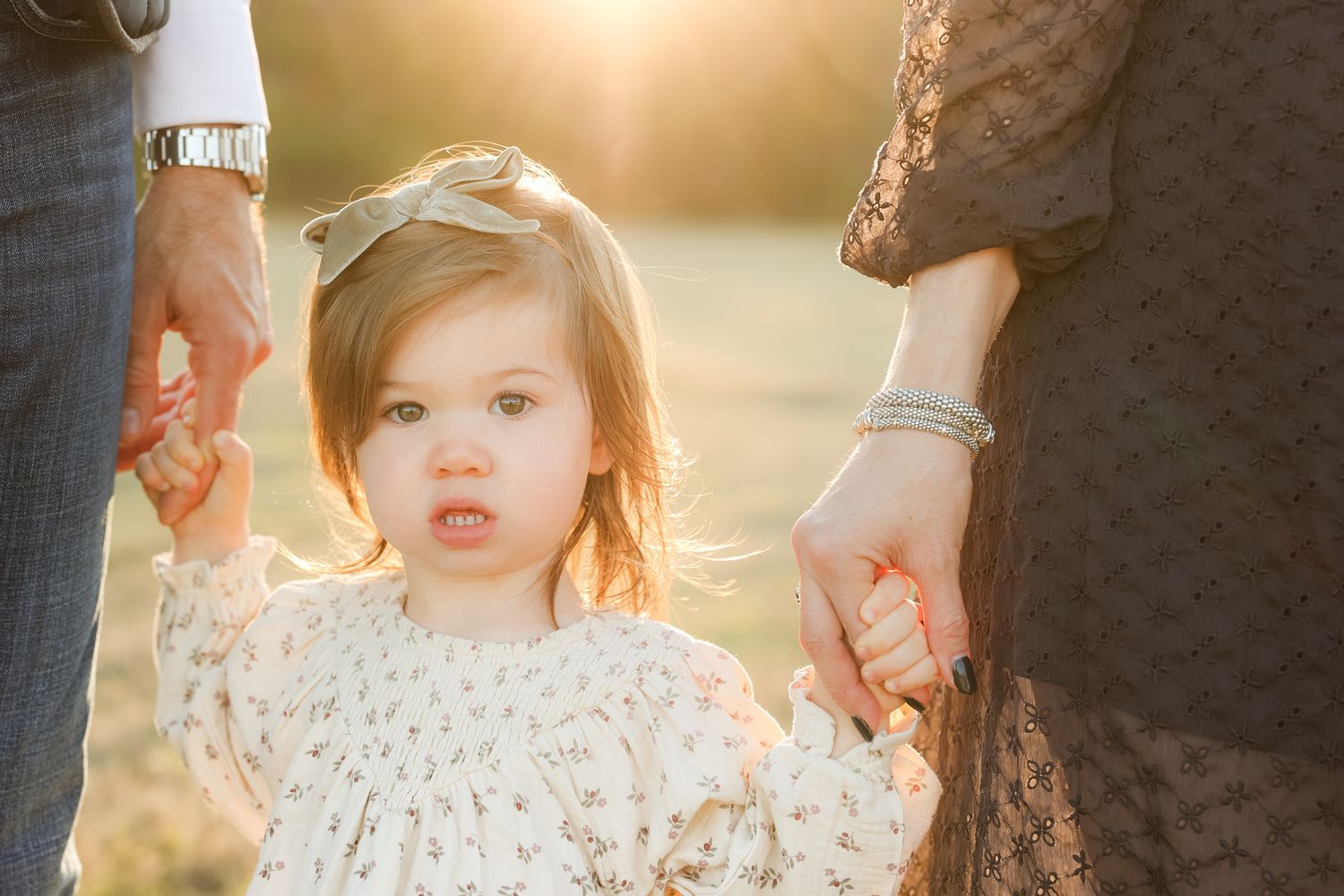 Backlit sunset portrait of small child in lace dress holding hands with adults.