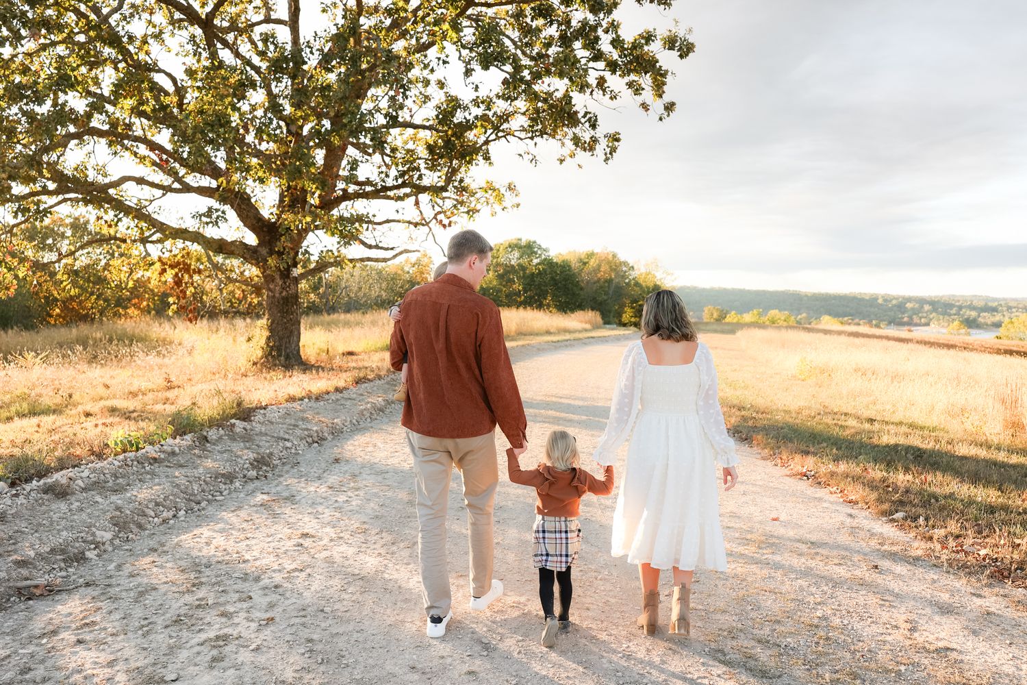 Family of three walking hand in hand down a dirt path at sunset with a large tree in the background.