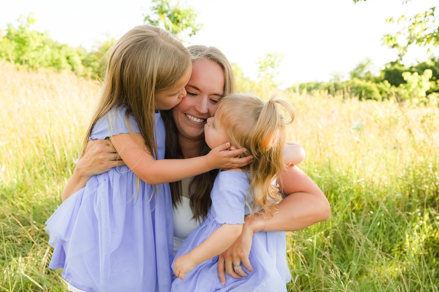 Three people in coordinating blue outfits sharing a loving embrace in a summer field.