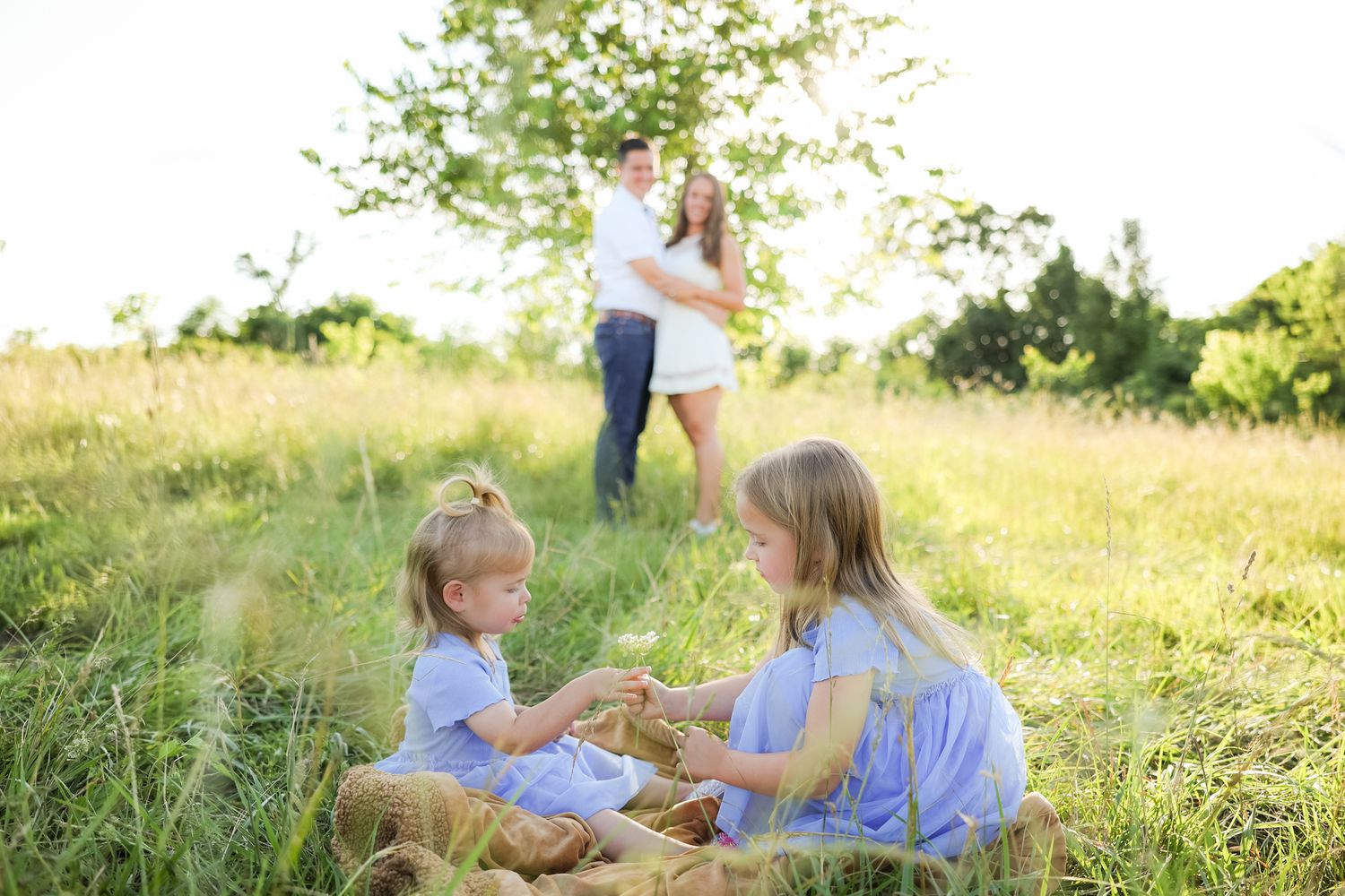 Children in blue dresses sitting in grass while adults embrace in background.