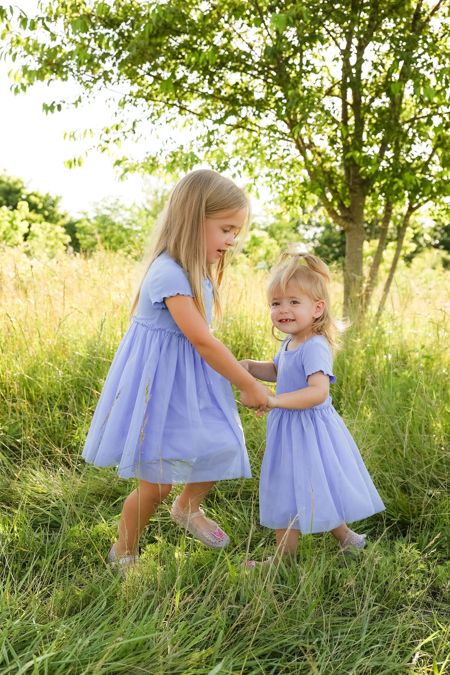 Two young girls in matching purple dresses playing together in a sunny meadow.