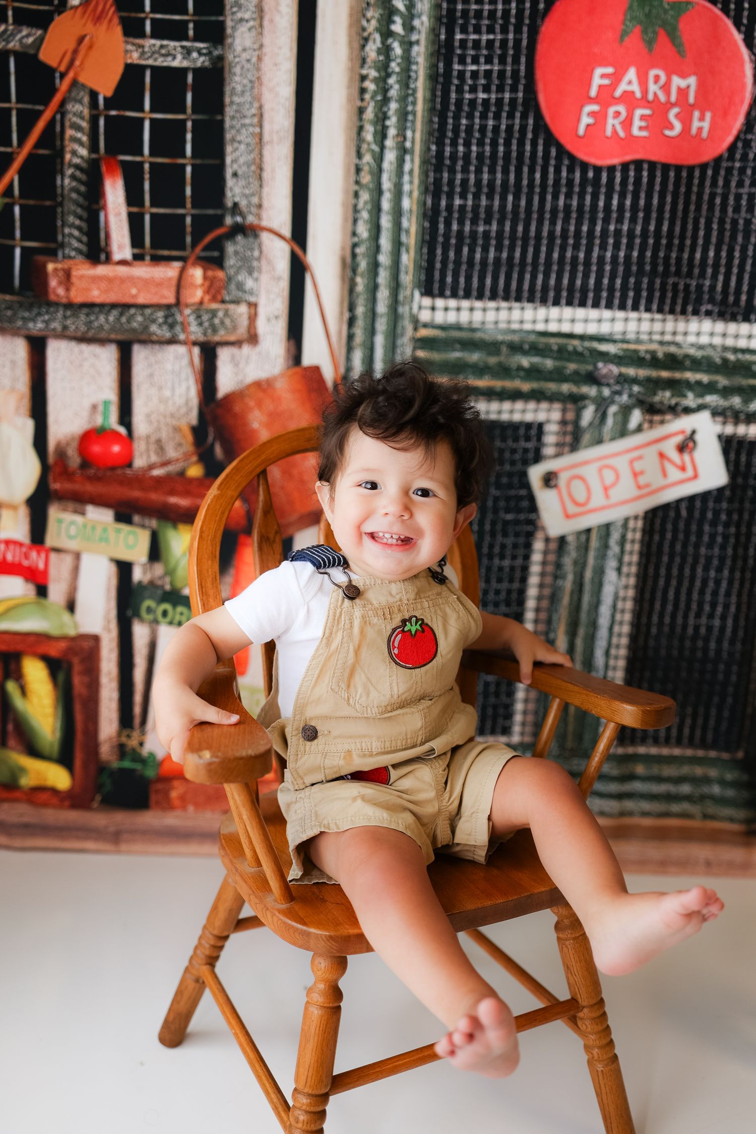 Toddler in overalls smiling while sitting on wooden chair with farm-fresh themed backdrop.