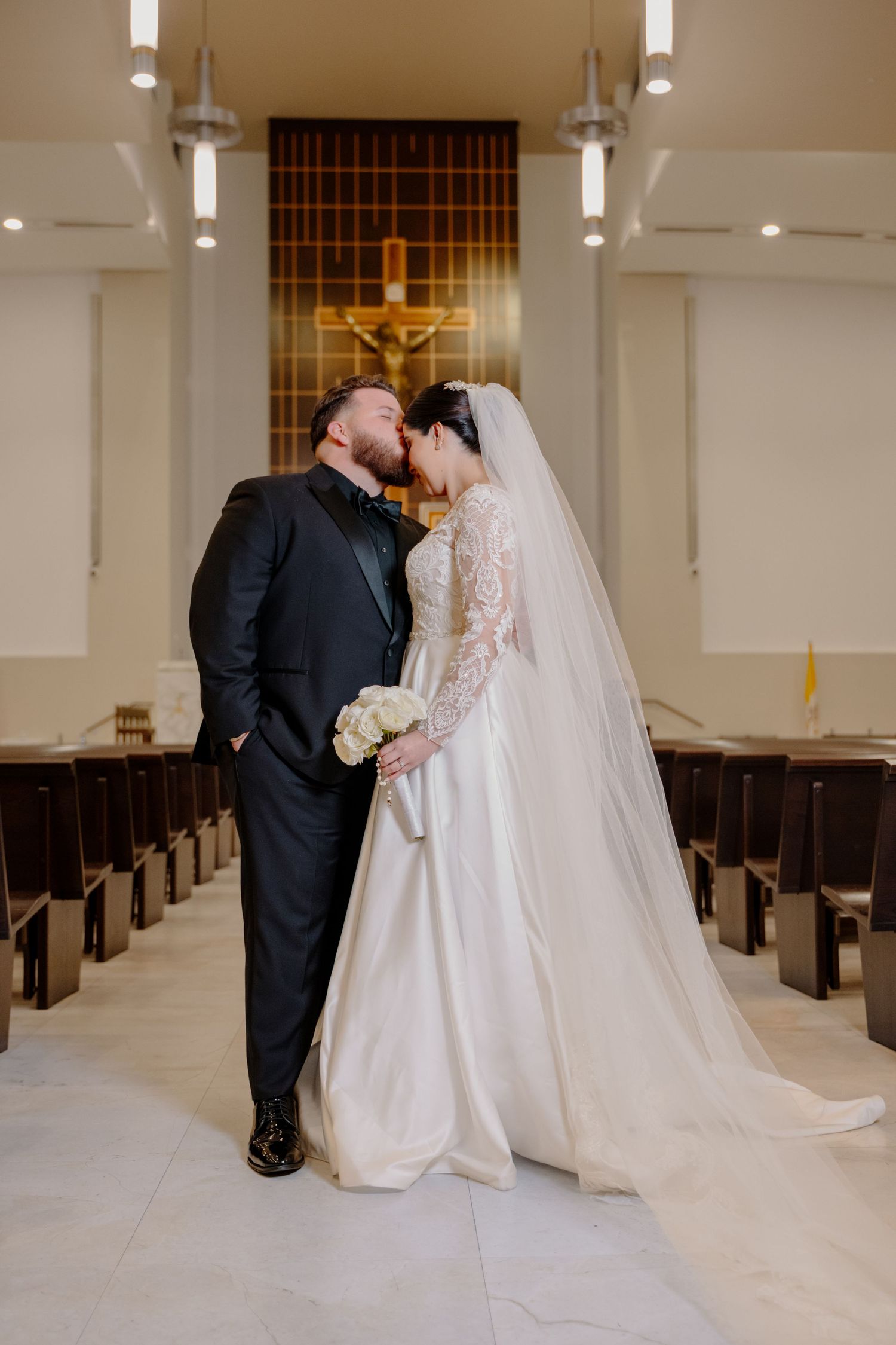 A bride and groom share an intimate moment in a church aisle with wooden pews and a crucifix in the background.