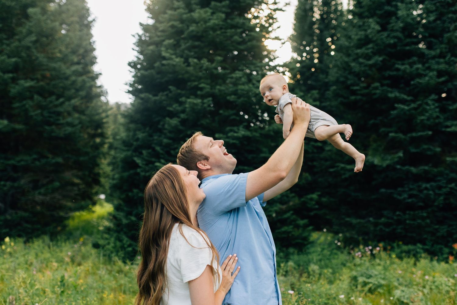 Young father lifts up his baby with mother looking lovingly at them in a Northern Utah mountain field