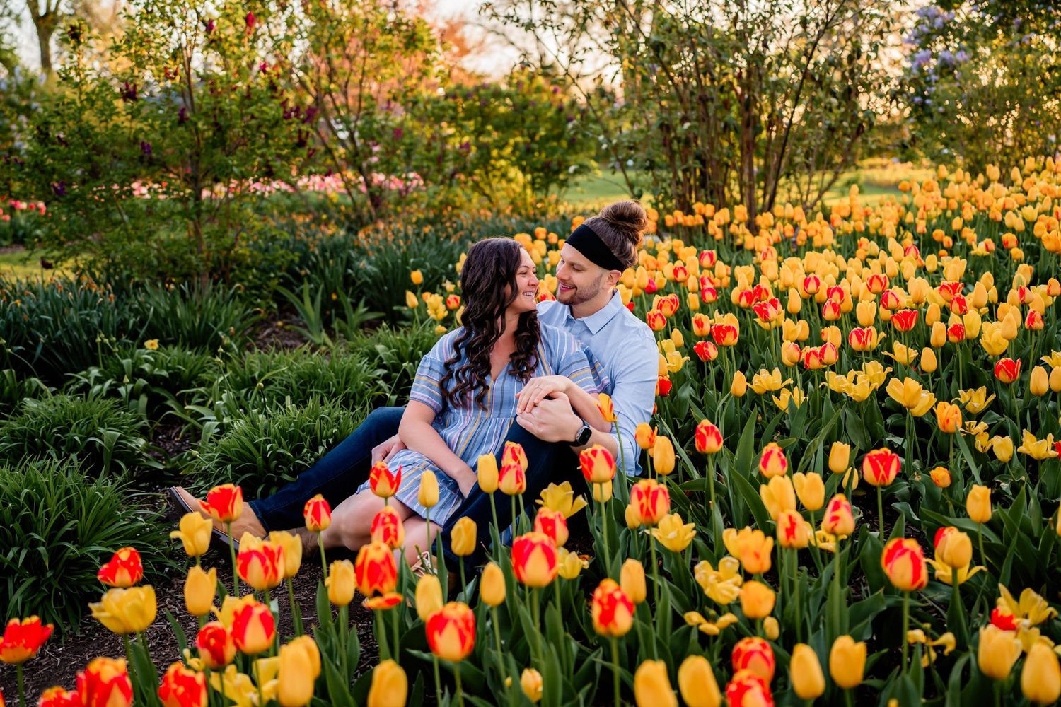 Two figures sit closely together in a vibrant garden filled with blooming orange and yellow tulips at sunset.