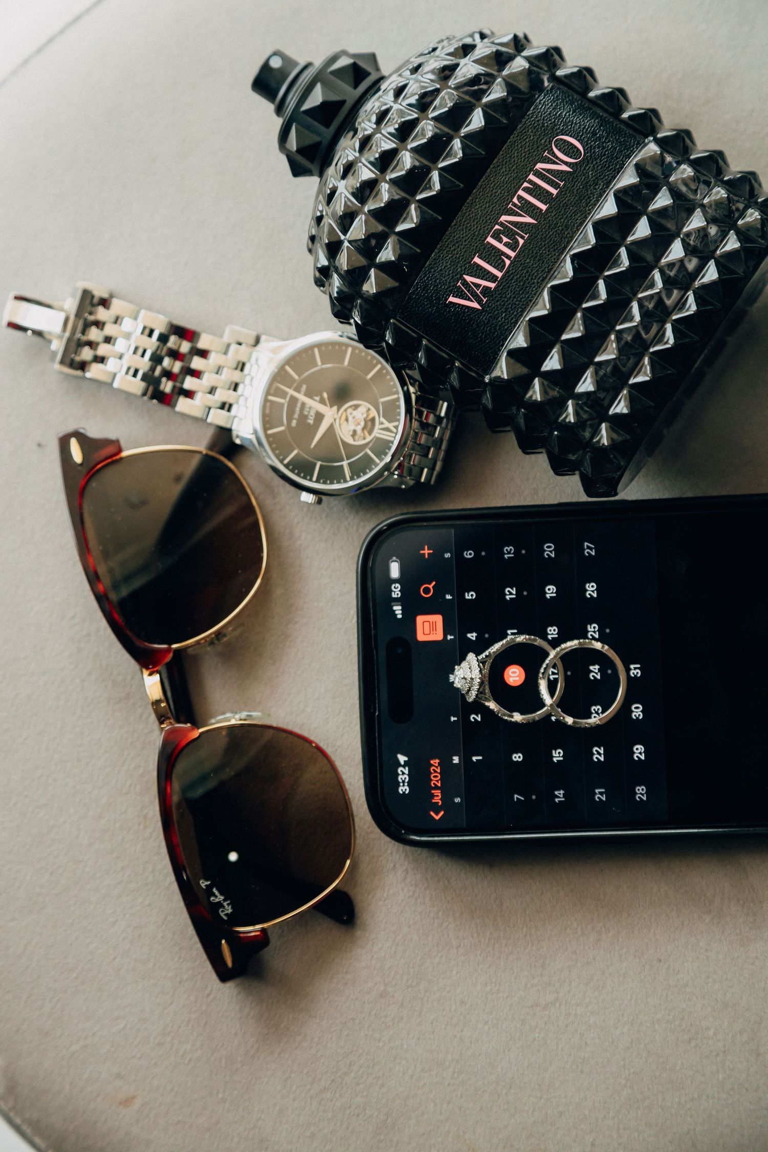 Stylish accessories including sunglasses, watch, and Valentino perfume bottle arranged on beige surface.