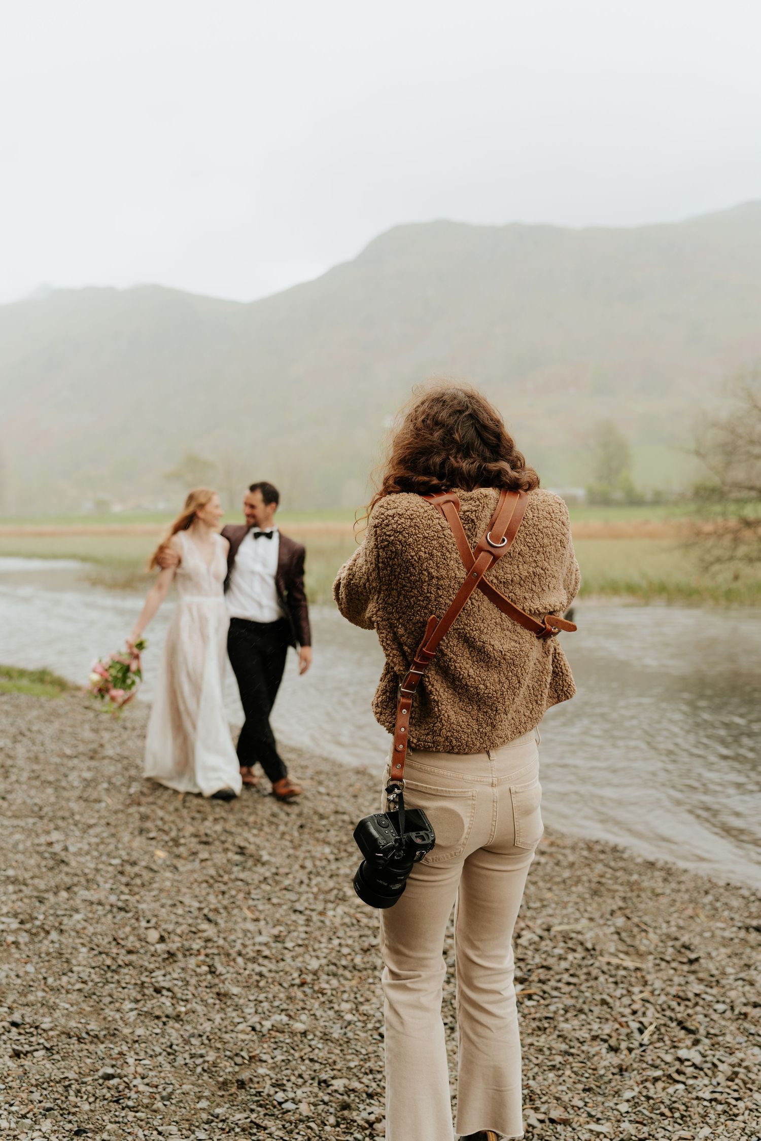 Behind-the-scenes shot of a photographer capturing a wedding couple walking along a lake shore.