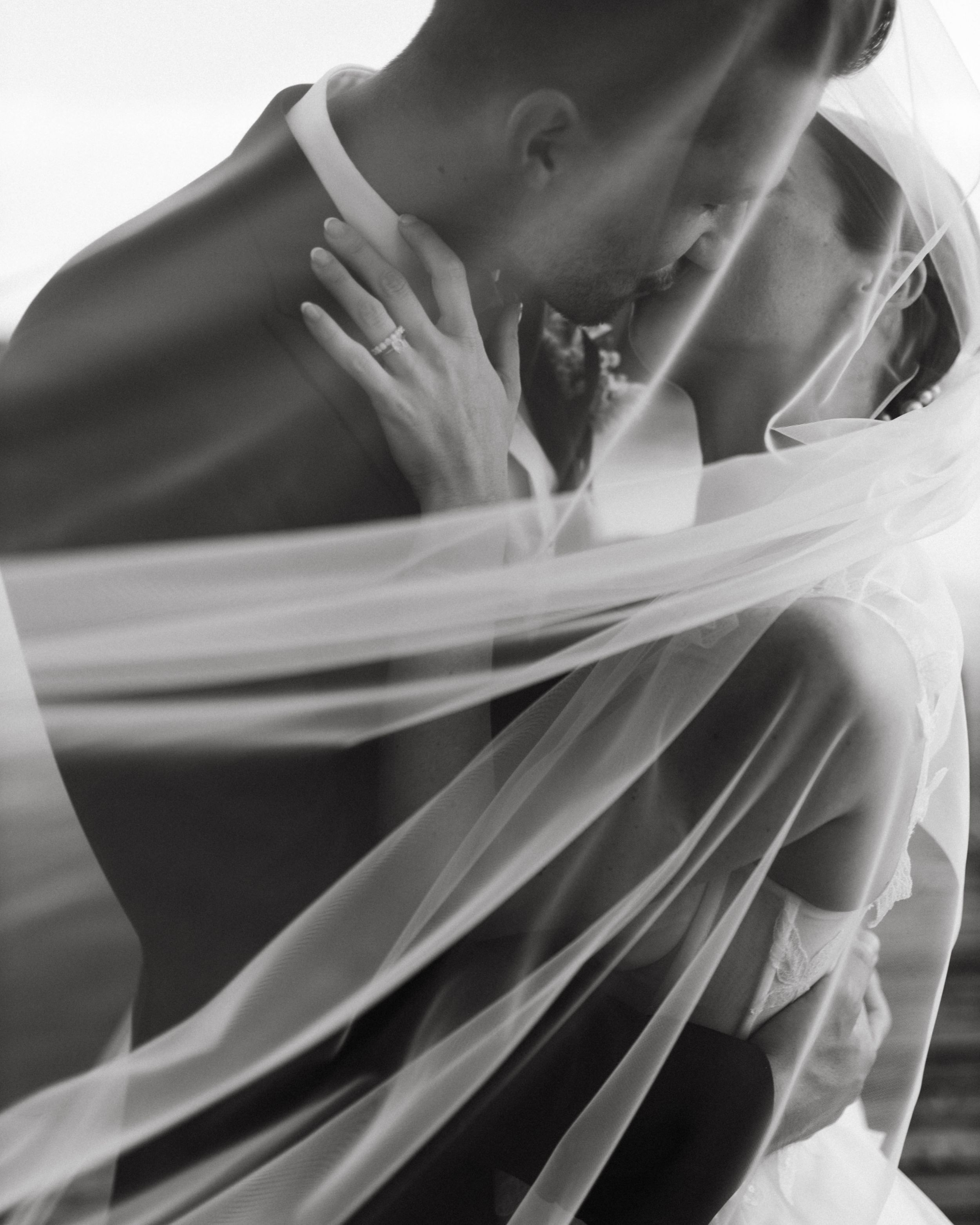 Artistic black and white wedding photo with a flowing veil creating an ethereal effect during an intimate moment.