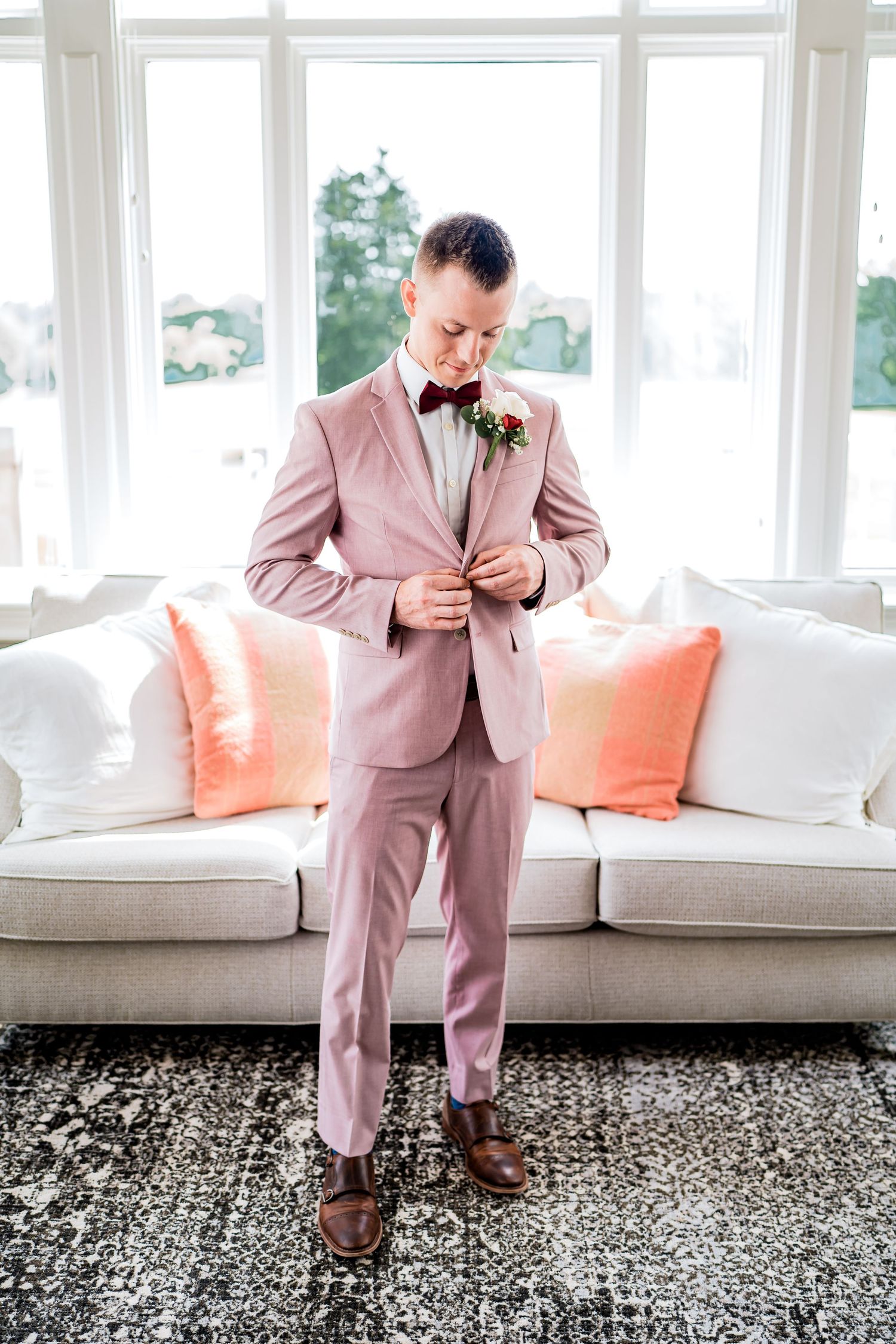 Groom in light pink suit and bowtie adjusts jacket while standing in front of white couch with coral accent pillows.