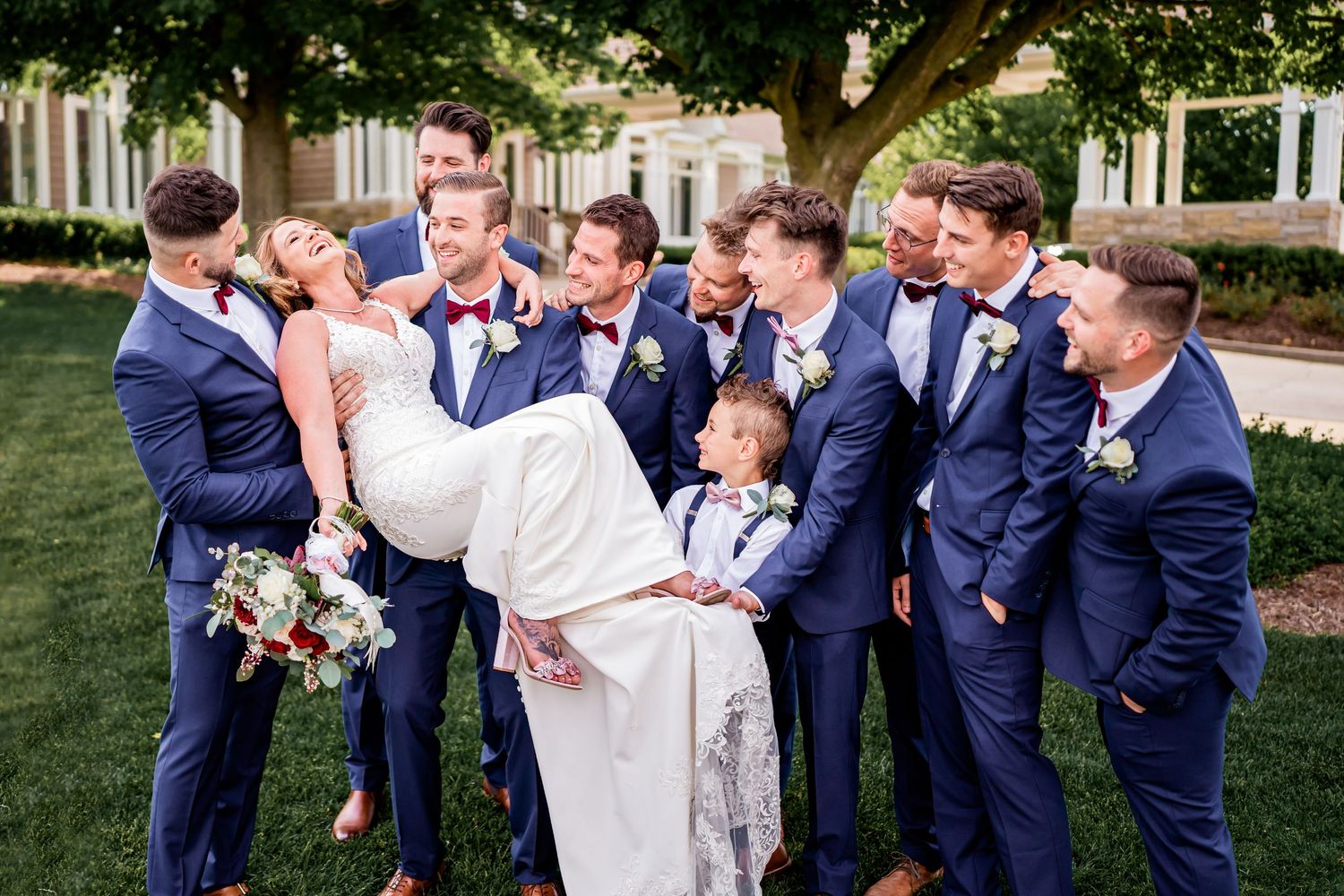 Wedding party in navy blue suits and white dress pose together playfully in a garden setting.