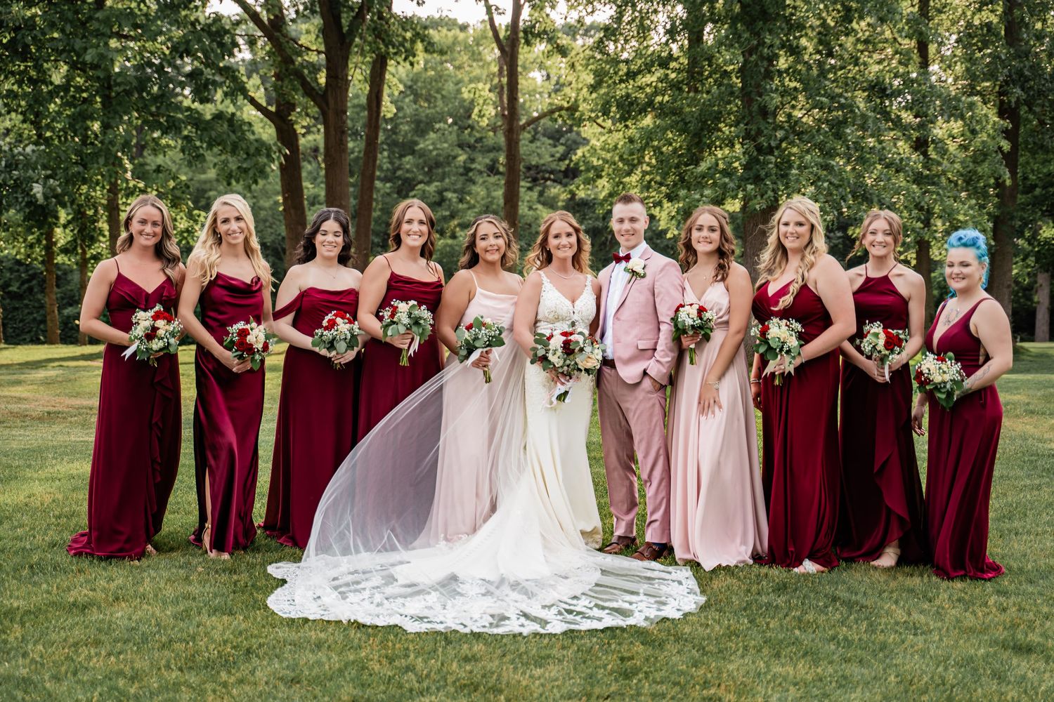 Wedding party in burgundy and blush bridesmaid dresses pose on green lawn during outdoor summer celebration.