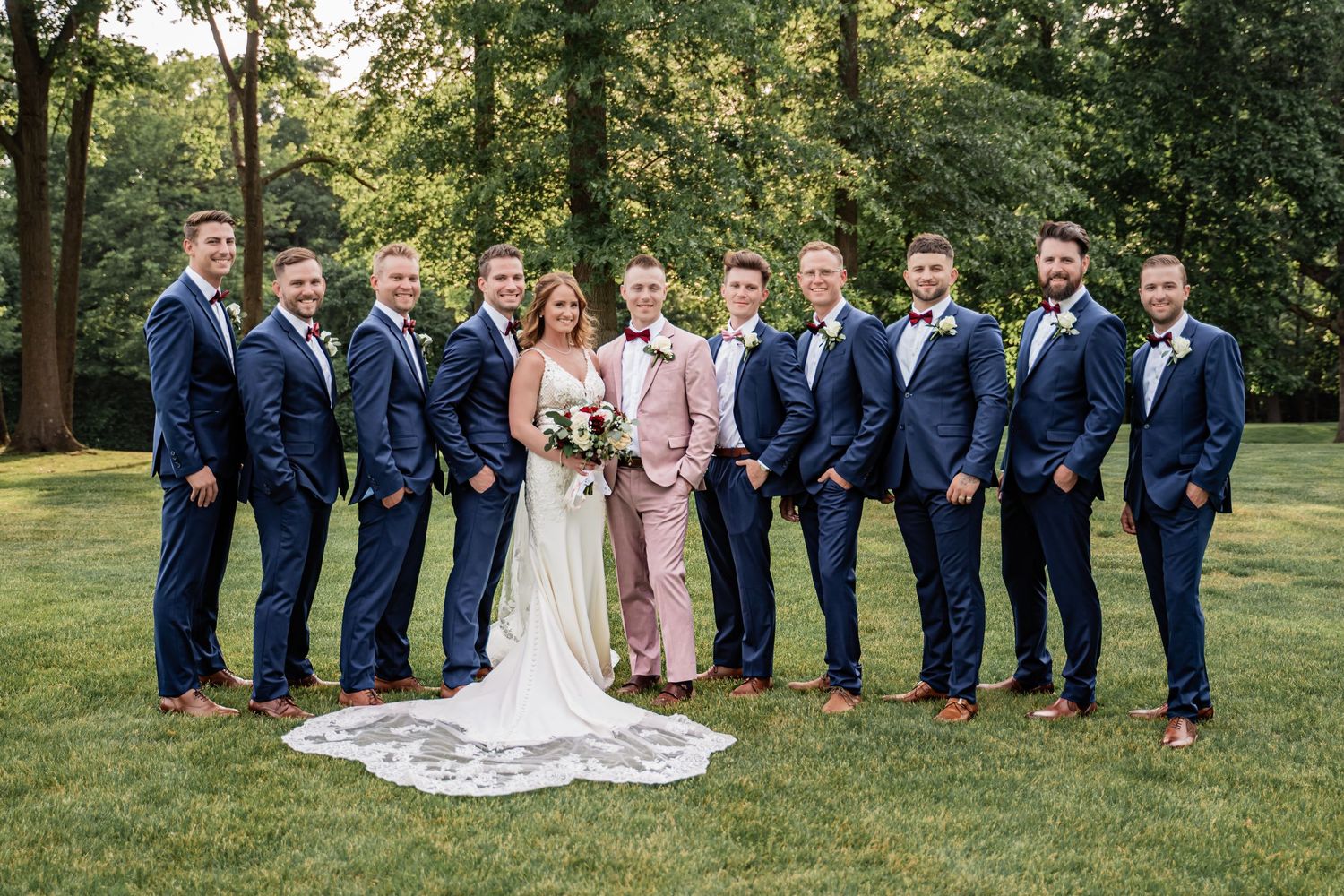 Wedding party in navy blue suits and pink dresses poses together on green lawn with trees in background.