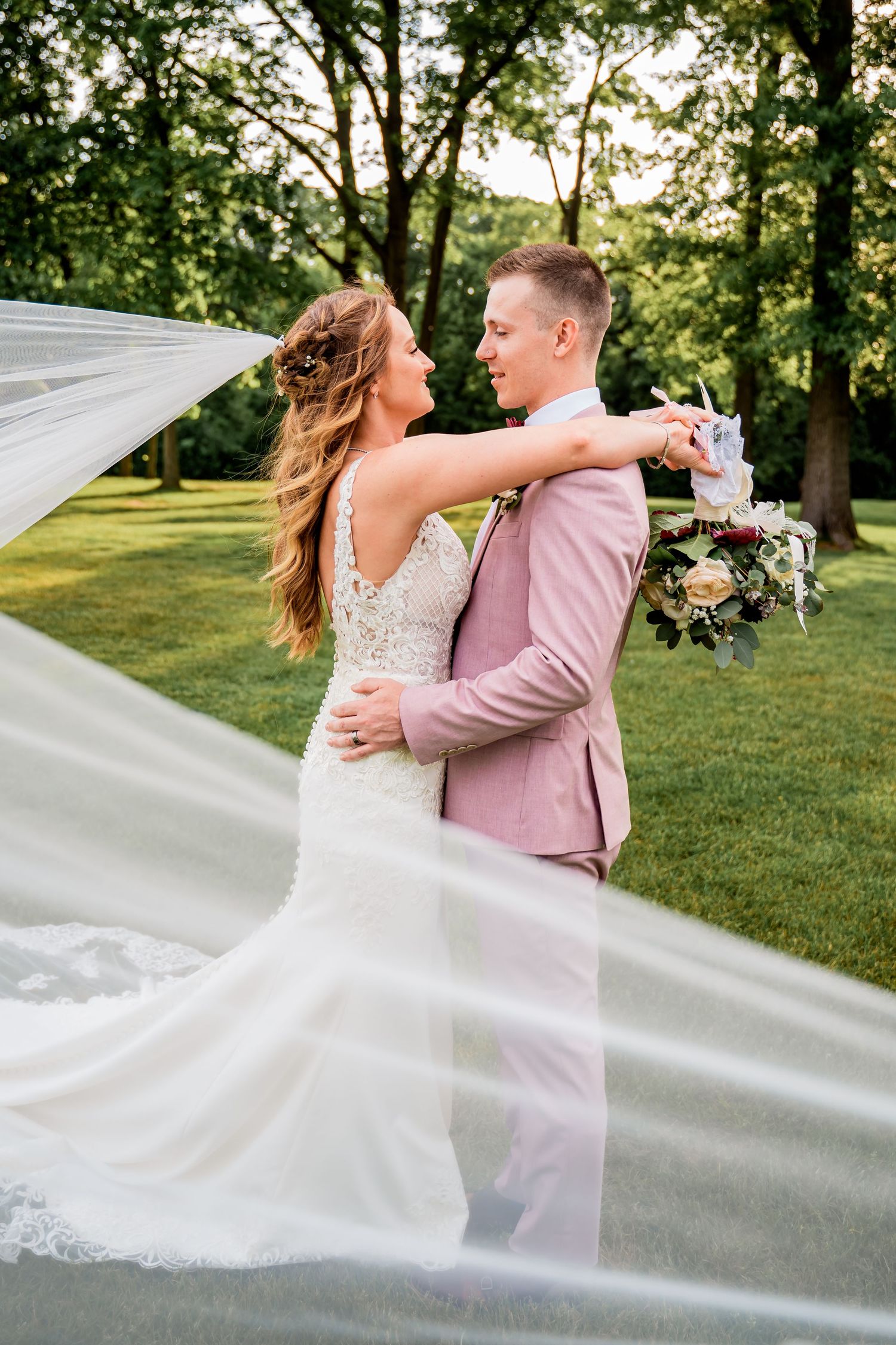 Romantic outdoor wedding moment captured with flowing white veil and mauve suit.